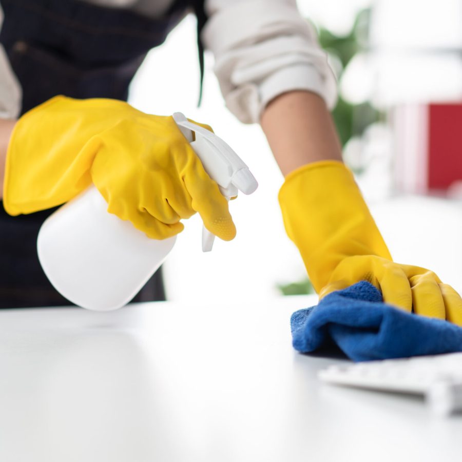 Housewife in apron wearing gloves to spraying hygiene spray on computer and using microfiber fabric to wiping cleanup the table while working and cleaning furniture in the house.